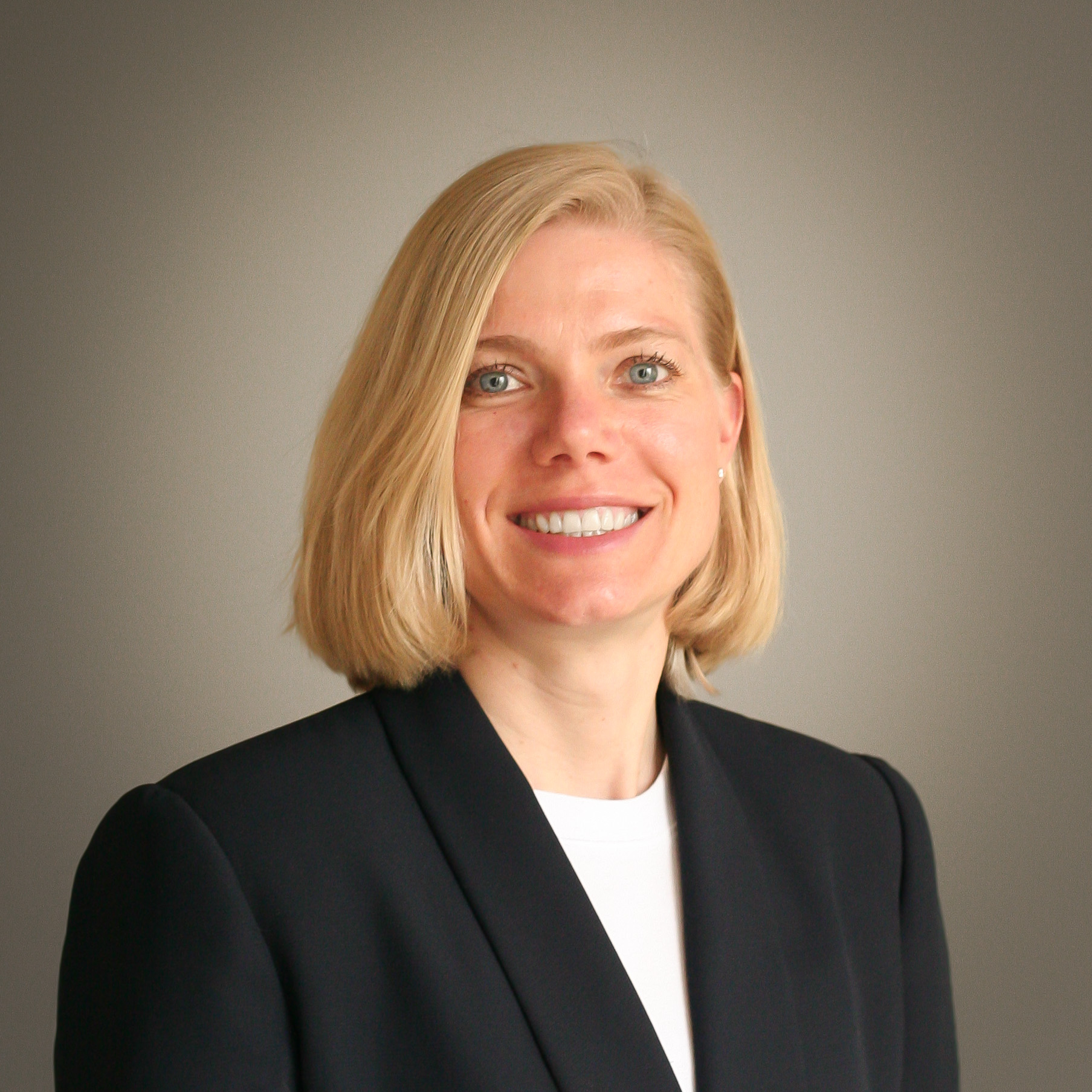 Square headshot.Professional portrait of Heather Albert, smiling, with short blonde hair, wearing a dark blazer over a white top, in front of a plain light background.